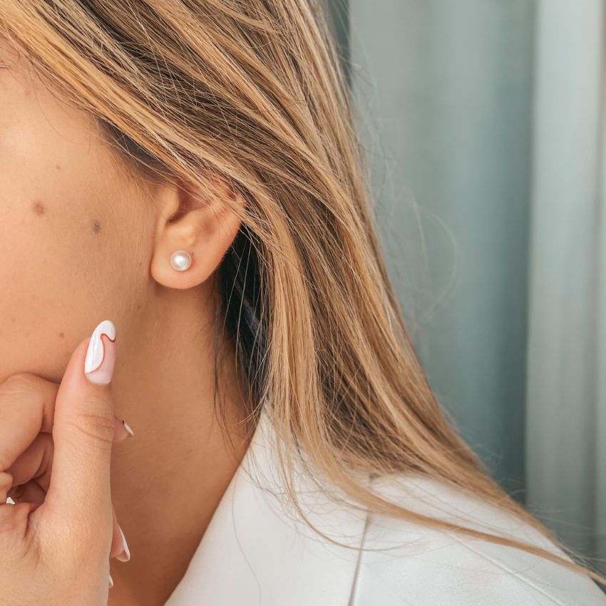 Close-up of a woman's ear wearing a silver pearl stud earring with styled nails touching her face.