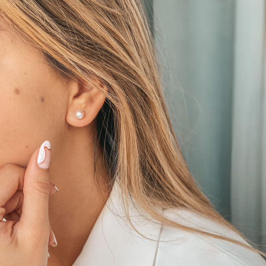 Close-up of a woman's ear wearing a silver pearl stud earring with styled nails touching her face.