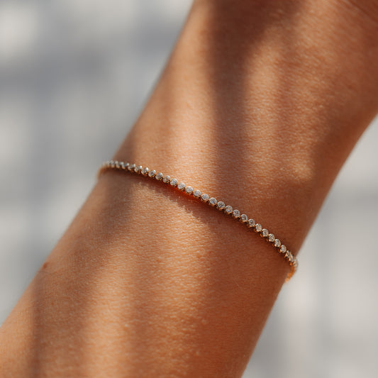 Detailní záběr na jemný tenisový náramek z 14K žlutého zlata s malými kulatými diamanty na zápěstí, osvětlený měkkým přirozeným světlem.