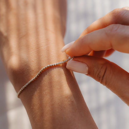 Close-up of a hand fastening a 14K yellow gold diamond tennis bracelet on a wrist in warm sunlight.