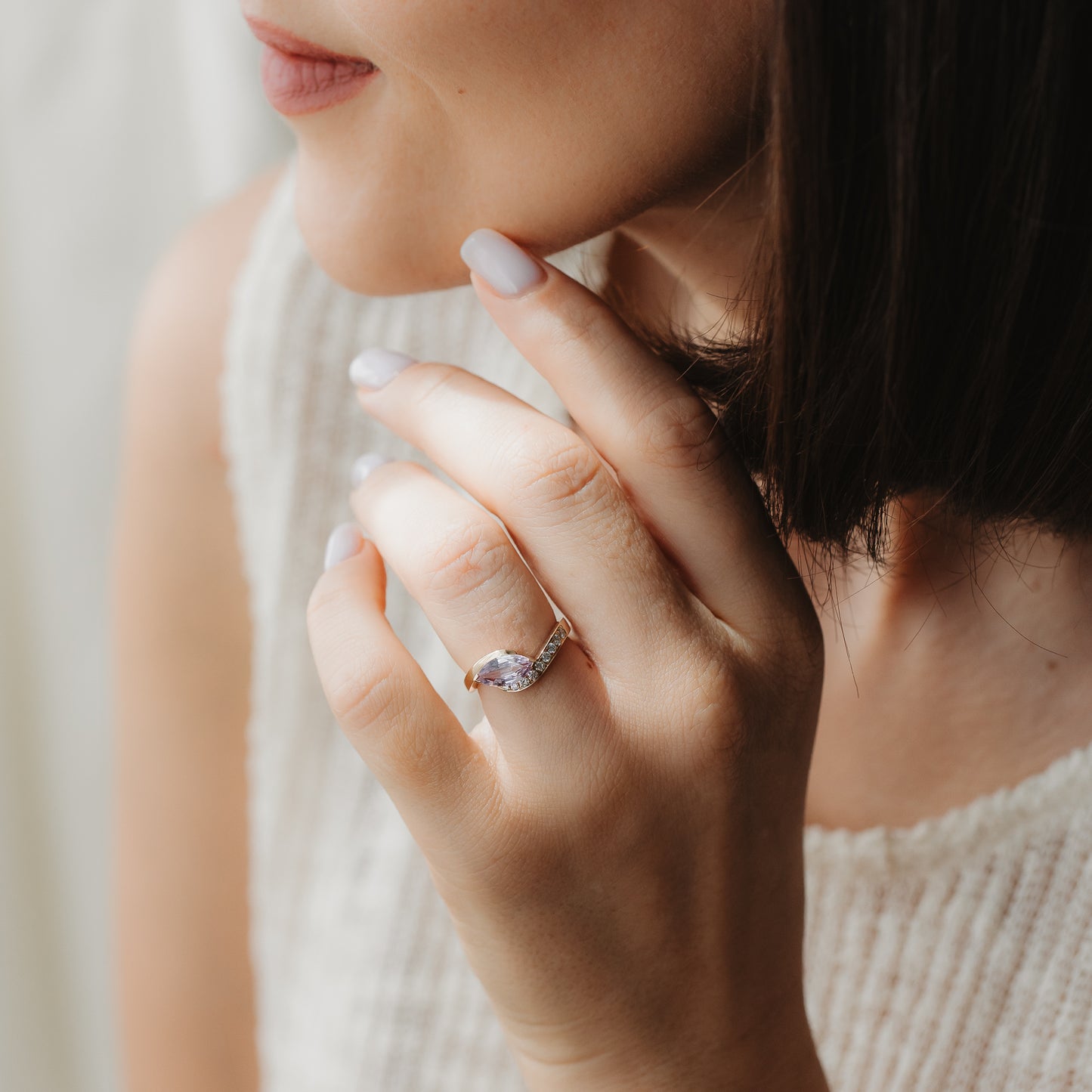 Close-up of a hand wearing a 14k yellow-gold marquise sapphire and diamond engagement ring, shown near the face in soft natural light.