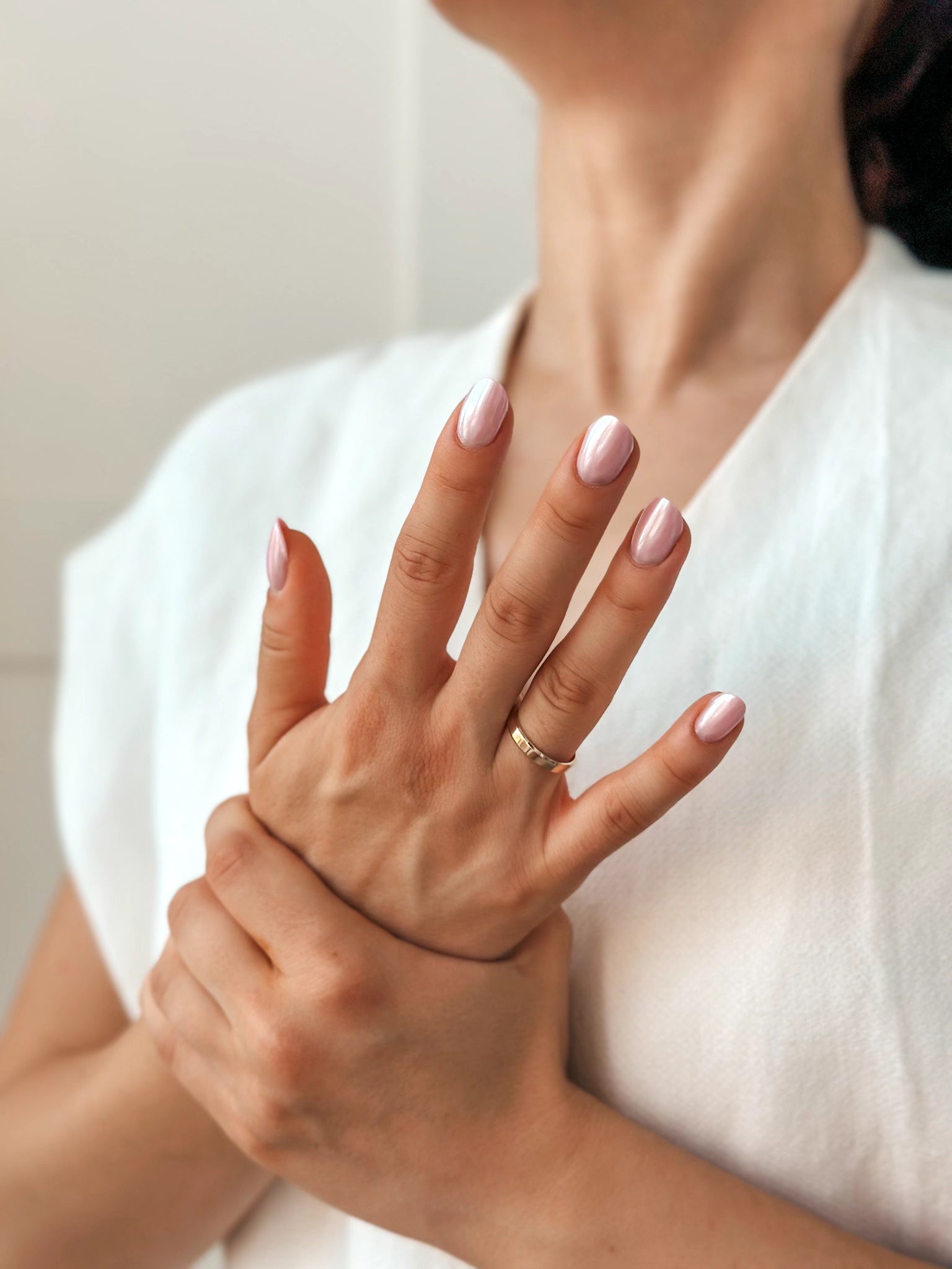 Close-up of a woman's hands with a pink nail polish and wearing a gold wedding band, against a light background.