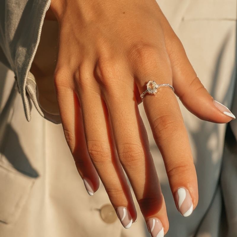 Close-up of a hand wearing a silver moonstone ring with a natural stone on a beige background.