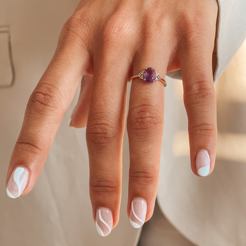 Close-up of a hand with a manicured nail wearing a radiant gold ring featuring an amethyst and diamonds.