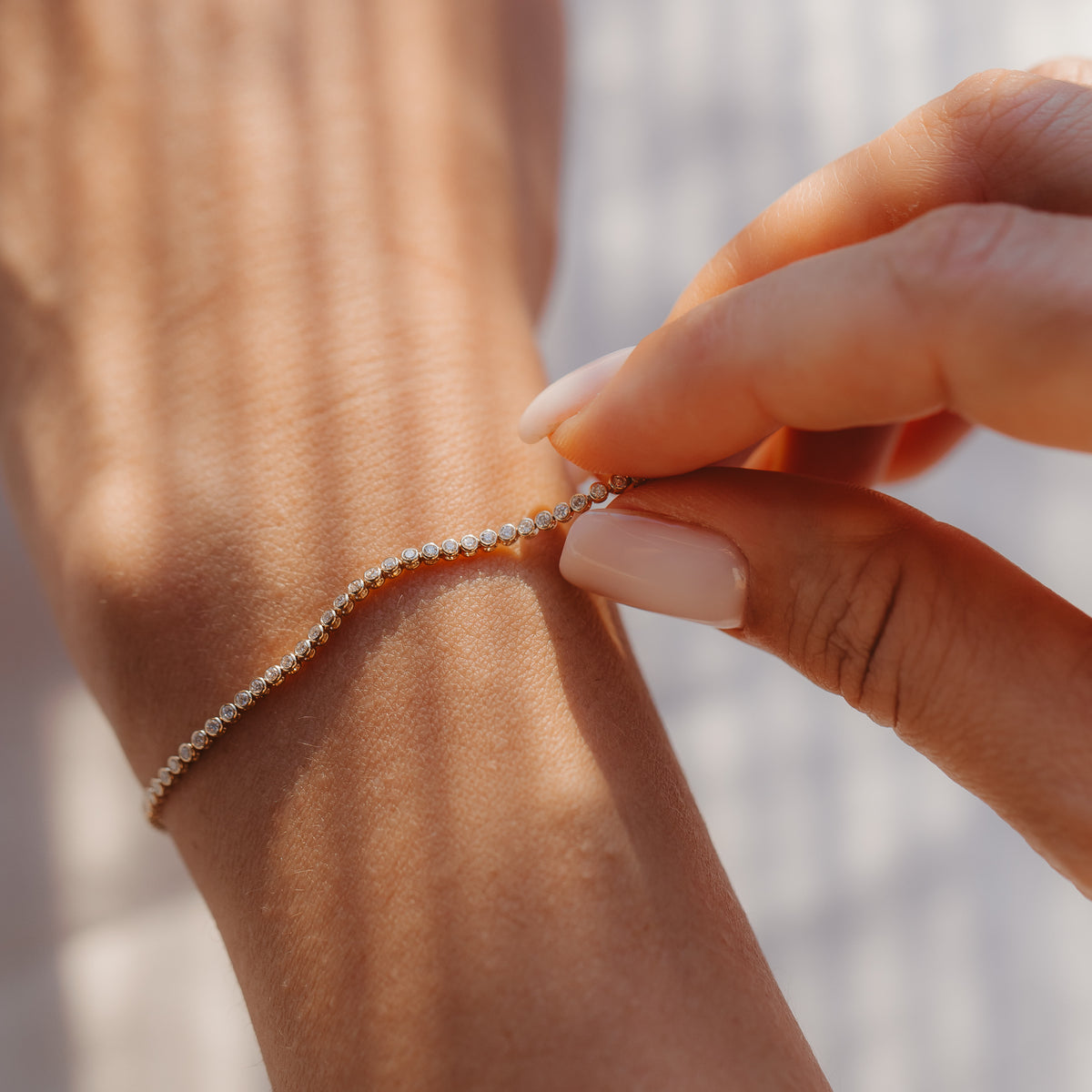 Detailní záběr ruky, která zapíná 14K tenisový náramek ze žlutého zlata s diamanty na zápěstí v jemném slunečním světle.