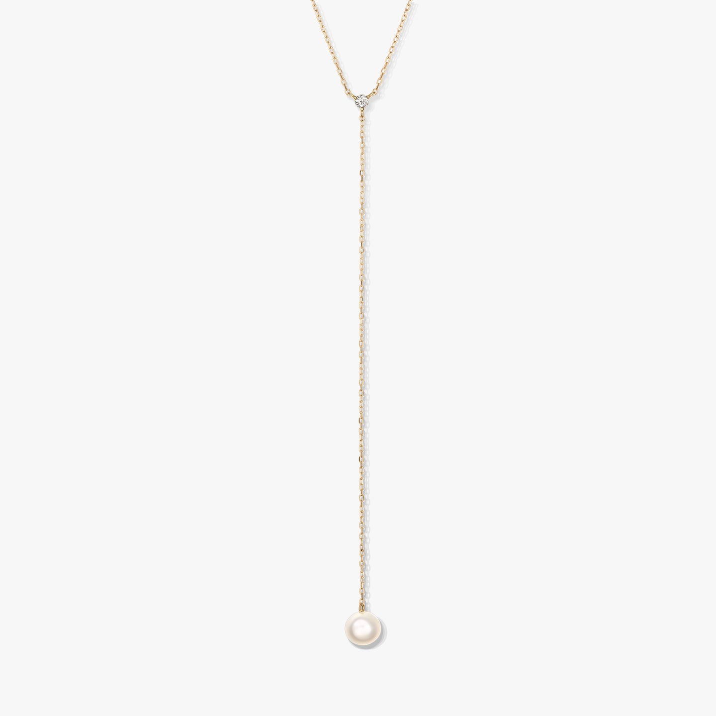 Minimalistický zlatý lariatový náhrdelník s jedinou kulatou bílou perlou ve tvaru kapky a malým doplňkovým kamínkem, vystředěný na bílém pozadí.