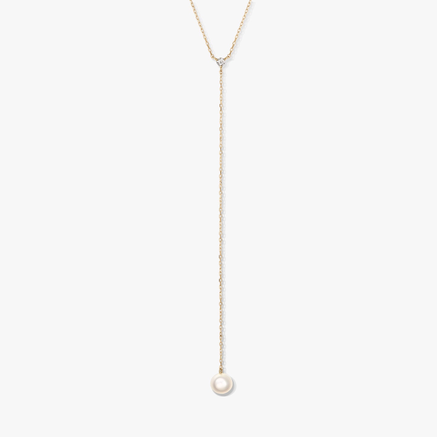 Minimalistický zlatý lariatový náhrdelník s jedinou kulatou bílou perlou ve tvaru kapky a malým doplňkovým kamínkem, vystředěný na bílém pozadí.