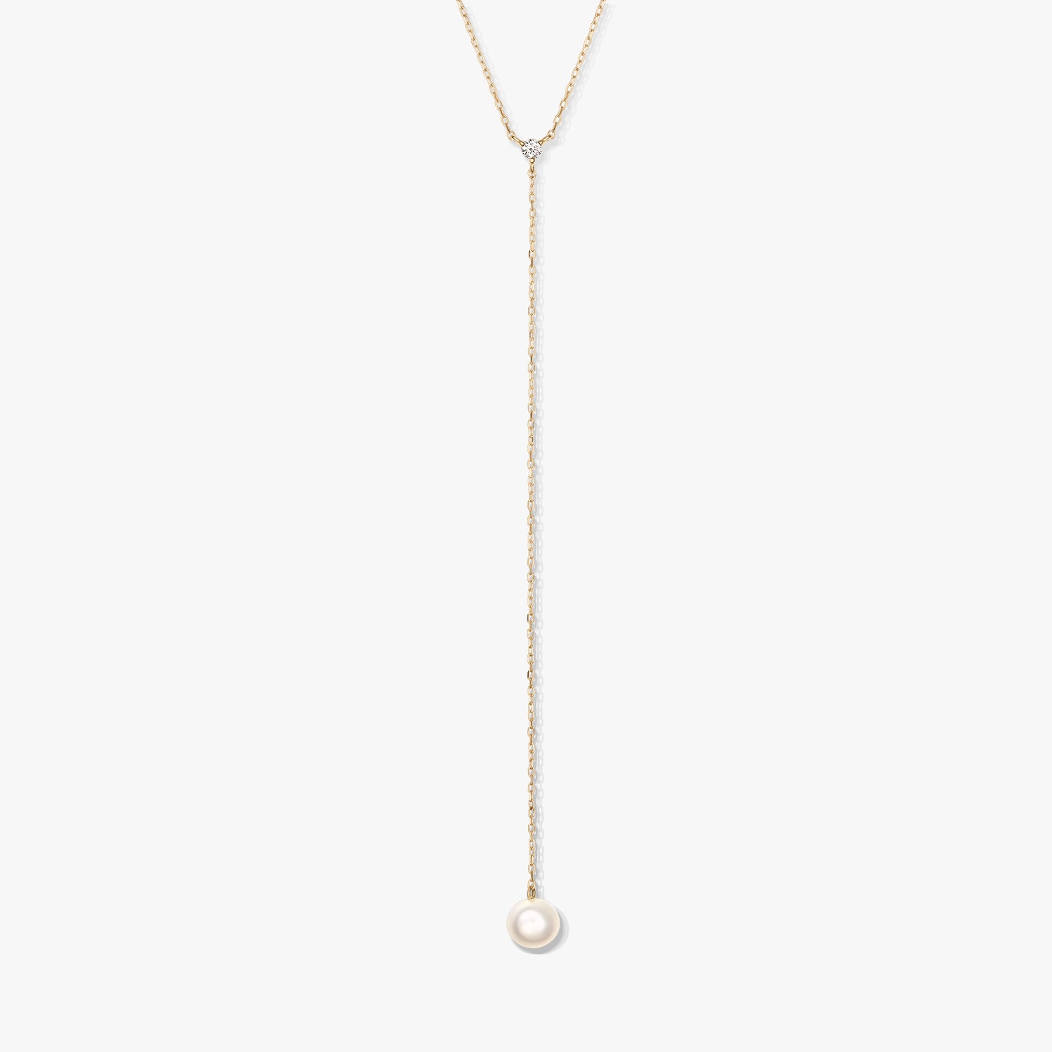 Minimalistický zlatý lariatový náhrdelník s jedinou kulatou bílou perlou ve tvaru kapky a malým doplňkovým kamínkem, vystředěný na bílém pozadí.