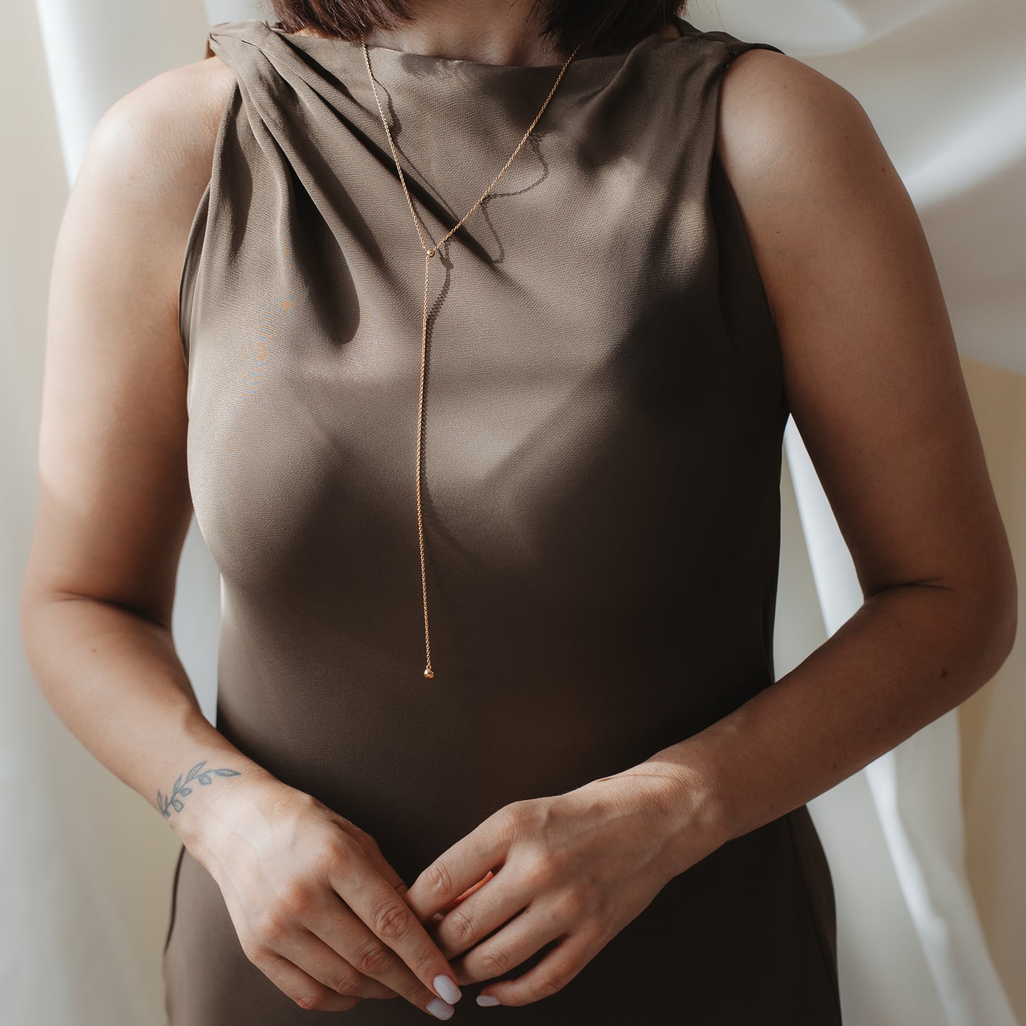 Close-up of a person wearing a gold‑plated 925 silver bola lariat necklace over a brown satin dress, hands gently clasped.