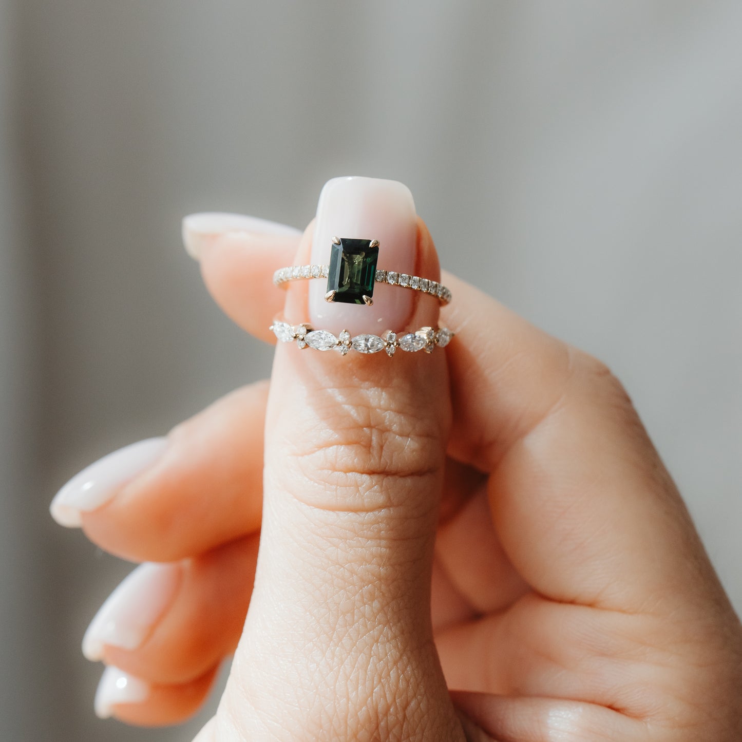 Detailní záběr ruky držící dva prsteny ze žlutého zlata: prsten Elysian se zeleným safírem ve smaragdovém brusu a s diamantovým pavé na obroučce, navrstvený nad prstenem Fiorelle s diamanty brusu markýza, na jemném šedém pozadí.