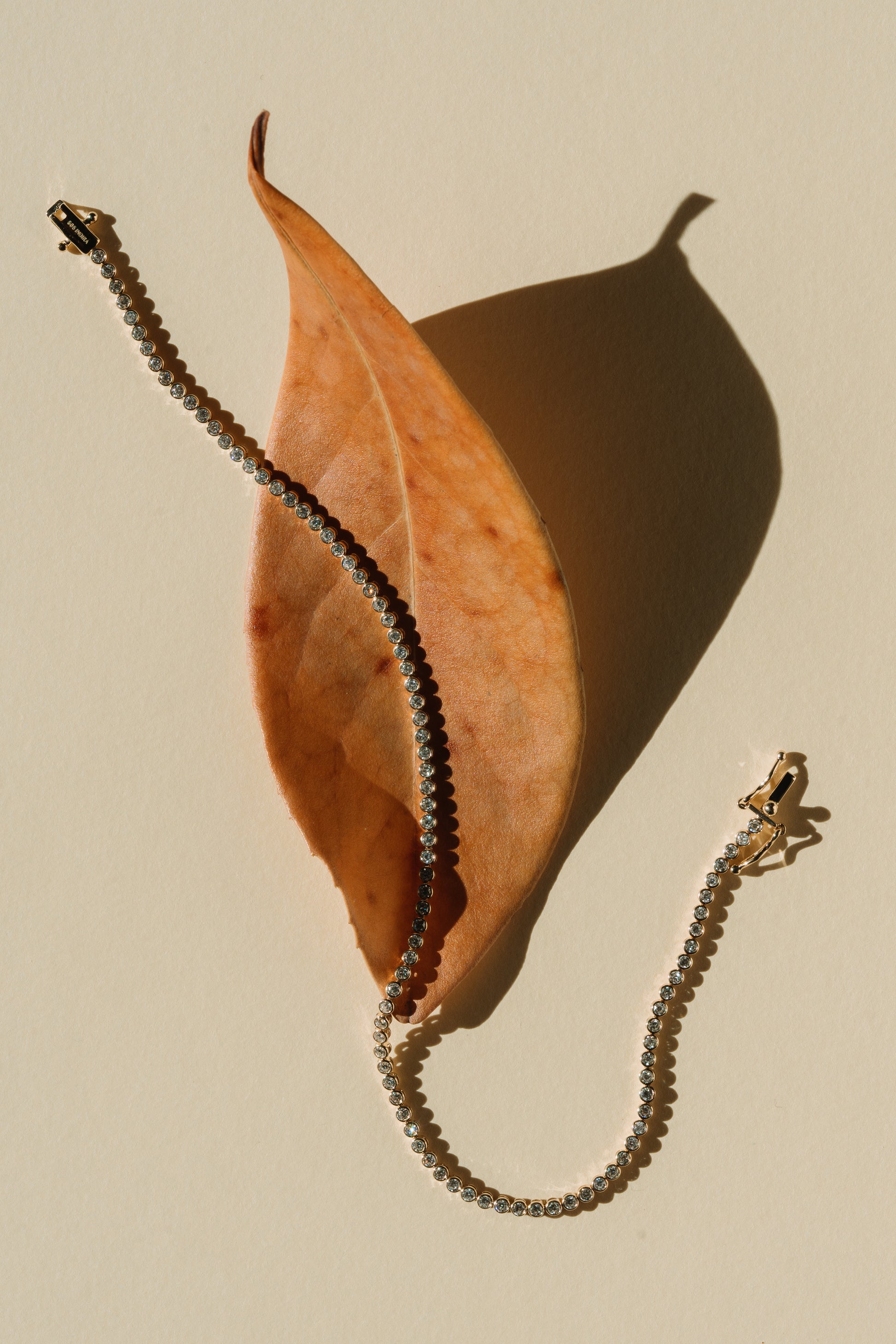 A gold tennis bracelet with diamonds resting on an autumn leaf against a beige background.