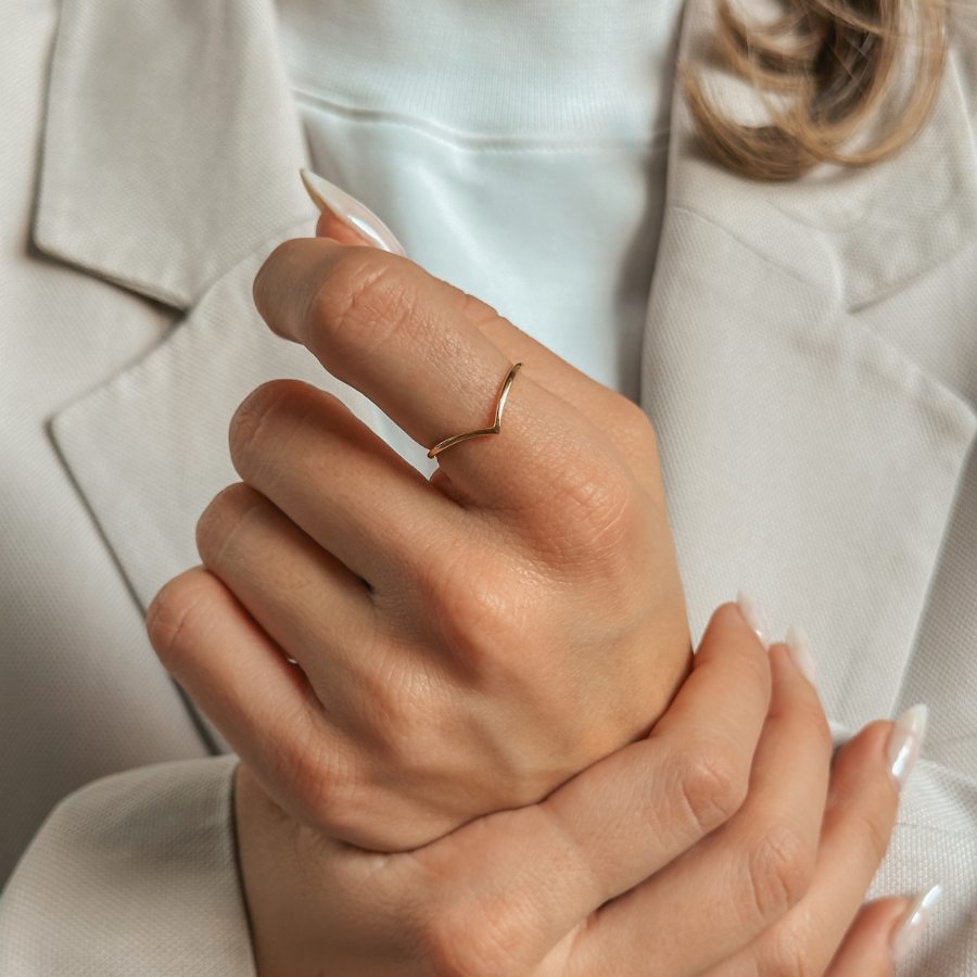 Detail ruky s jednoduchým prstenem ve tvaru V, pozlaceným stříbrem, na pozadí s neutrálním oblečením.
