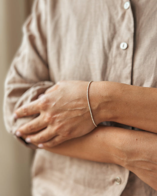 Close-up of an arm wearing a delicate bracelet, with hands crossed, dressed in a linen shirt.