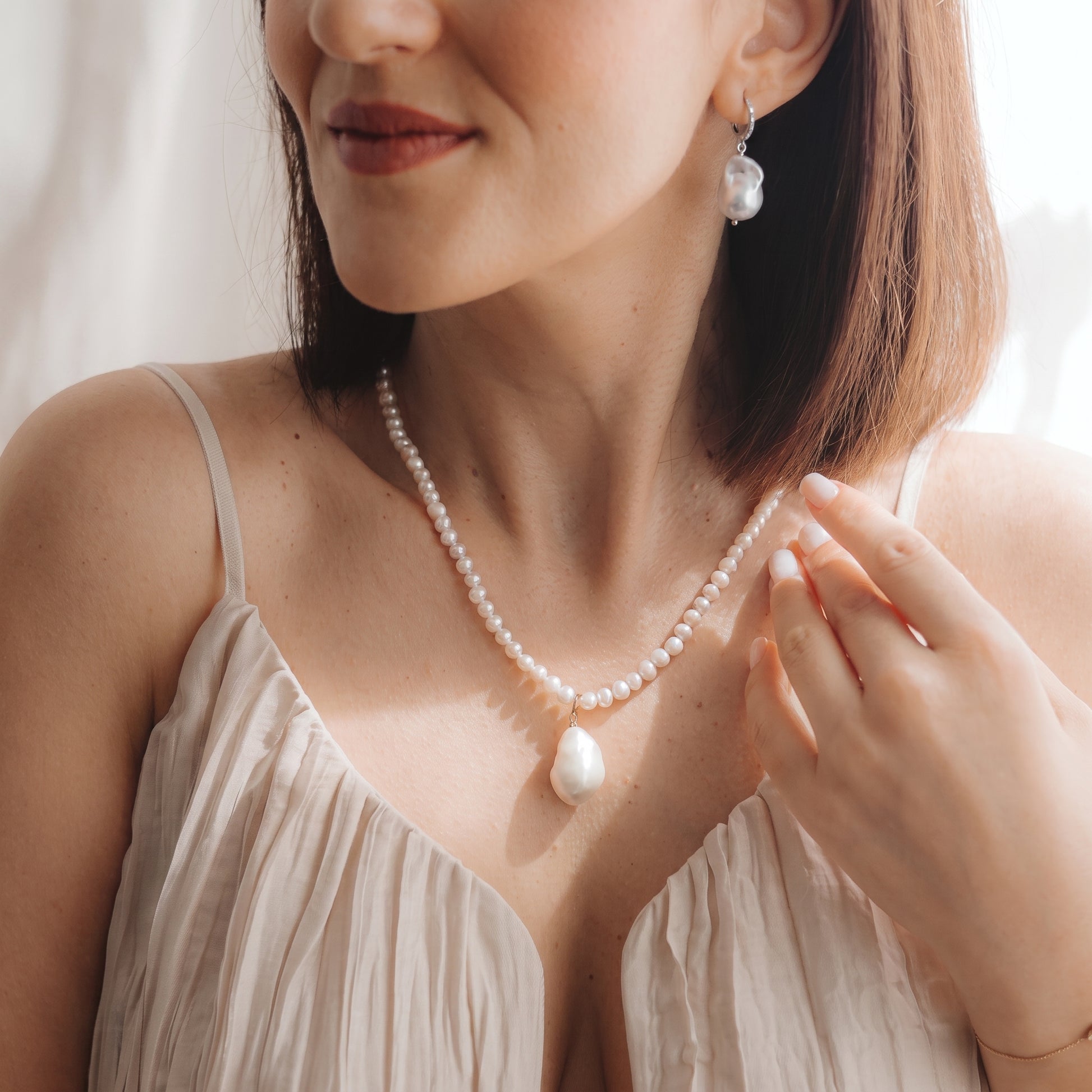 Close-up of a model wearing a rhodium-plated 925 silver baroque pearl drop earring with a small zirconia detail and a matching pearl necklace, hand near the collarbone in soft natural light.