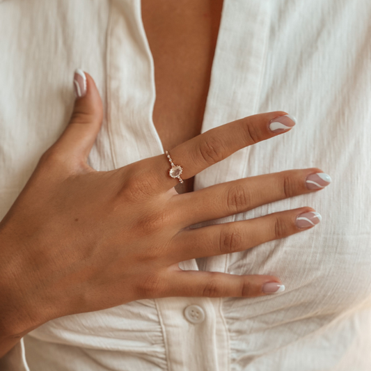 Close-up of a hand with a rose gold ring featuring a moonstone on a woman's finger with manicured nails and a white blouse.