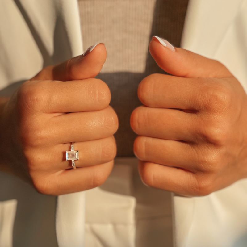 Blízký záběr rukou nosících stříbrný prsten Evie s měsíčním kamenem, bílé oblečení, se sepnutými palci.