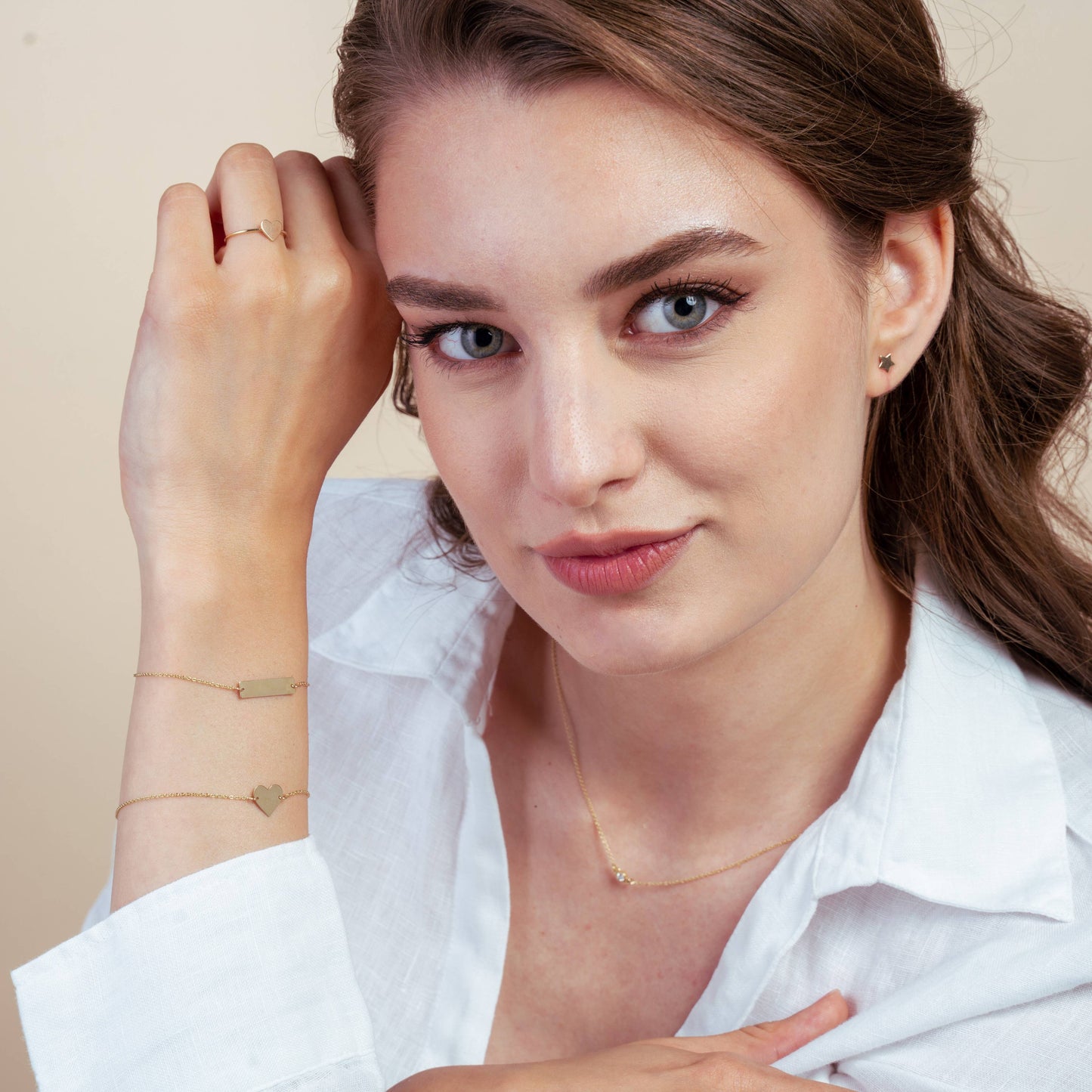 Woman wearing a white shirt, showcasing gold jewelry including a heart bracelet, small earrings, and a delicate necklace in a studio setting.