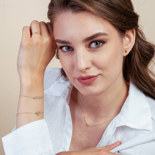 Woman wearing a white shirt, showcasing gold jewelry including a heart bracelet, small earrings, and a delicate necklace in a studio setting.
