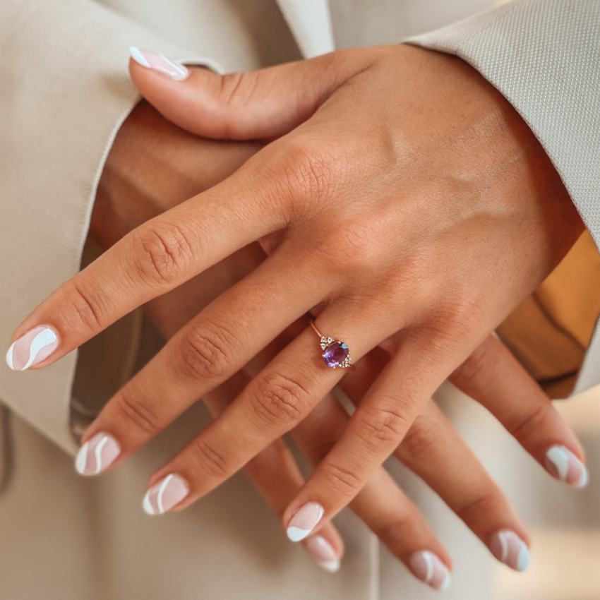 A close-up of a hand wearing a gold ring with an amethyst gemstone, surrounded by diamonds, on manicured fingers.